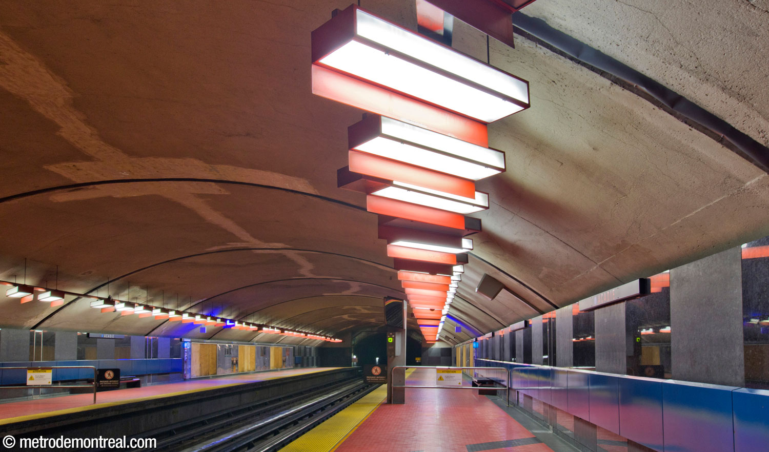 View of the Saint-Michel platform, looking toward the closed-off north end of the platforms. Above, the platform lighting consists of standard metro light fixtures but painted red, set at the station's signature 45-degree angle, and supplied with shades that throw the light dramatically onto the walls and floor.