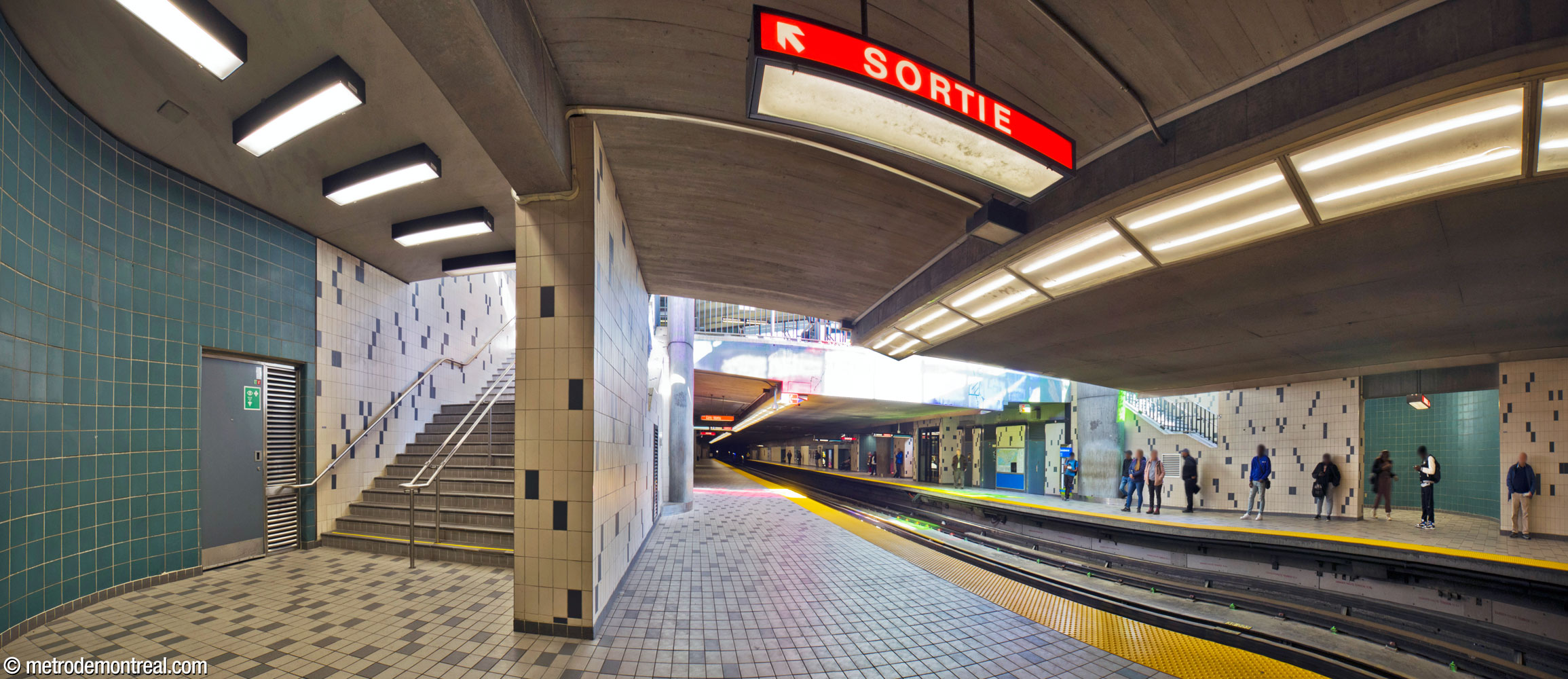 The main stairwells from the ticket hall to the platforms pass through the open volume at the west end of the headhouse, where the stained-glass window admits coloured light to the platforms. They pass behind the large columns supporting the roof and behind a section of platform wall before opening onto the platforms. This view looks east from the Côte-Vertu platform.