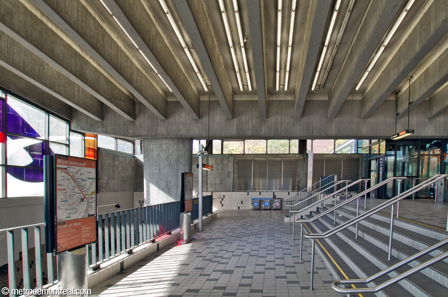 The lower level of the ticket hall at its western end. The wide staircase at right leads down from the gatelines; the parapet at left overlooks the platforms. The ends of this lower level lead to the staircases down to the platforms; this view looks toward the concrete northern wall of the headhouse and the heads of the staircases to the Côte-Vertu platform.