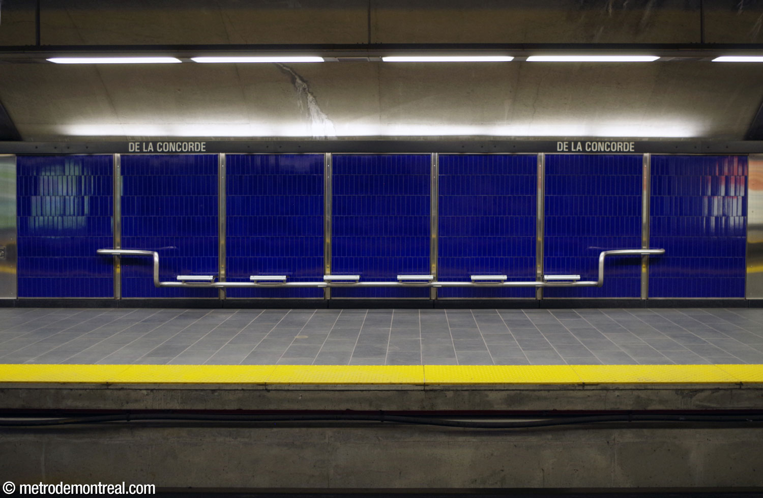 L'un des bancs en acier inoxydable, qui ressemblent à ceux des autres stations lavalloises, sur le fond bleu saphir du mur du quai.