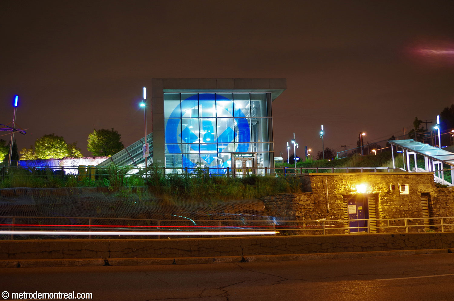 Le mur-rideau en verre sur la façade de l'édicule rayonne la nuit, mettant en valeur le logo du métro qu'il arbore.