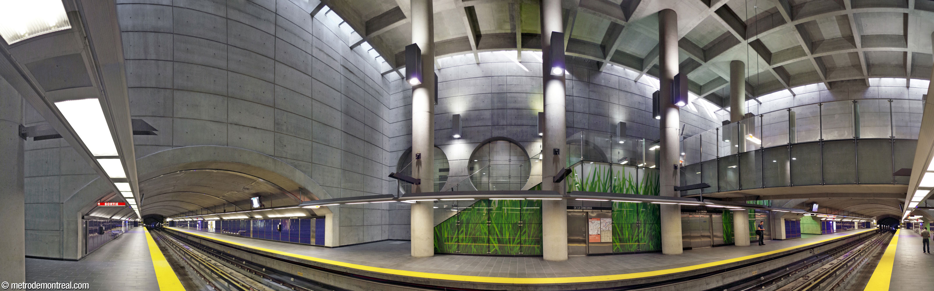 Panorama de la nef à partir du quai Côte-Vertu. Les extrémités des quais, construites en tunnel, se projettent du grand volume, créant ainsi des ouvertures dans les murs hauts et plats en béton. Dans le grand volume central, de forme cubique, de hautes colonnes soutiennent le plafond à caissons, qui est entouré sur tout son pourtour par des puits de lumière. Des lampes éclairent tant le haut que le bas des colonnes et des murs. Au centre, un escalier mène vers la passerelle (droite). Le côté de l'escalier est égayé par une grande murale verte et blanche, une photographie en grand plan de brins d'herbe. Derrière et en haut de cet escalier, une suite de trois grandes fenêtres rondes s'ouvre vers le couloir entre la billetterie et l'ascenseur vers la surface. Presque toutes les surfaces sont en béton brut, hormis les murales photographiques et les murs bleu saphir des quais.