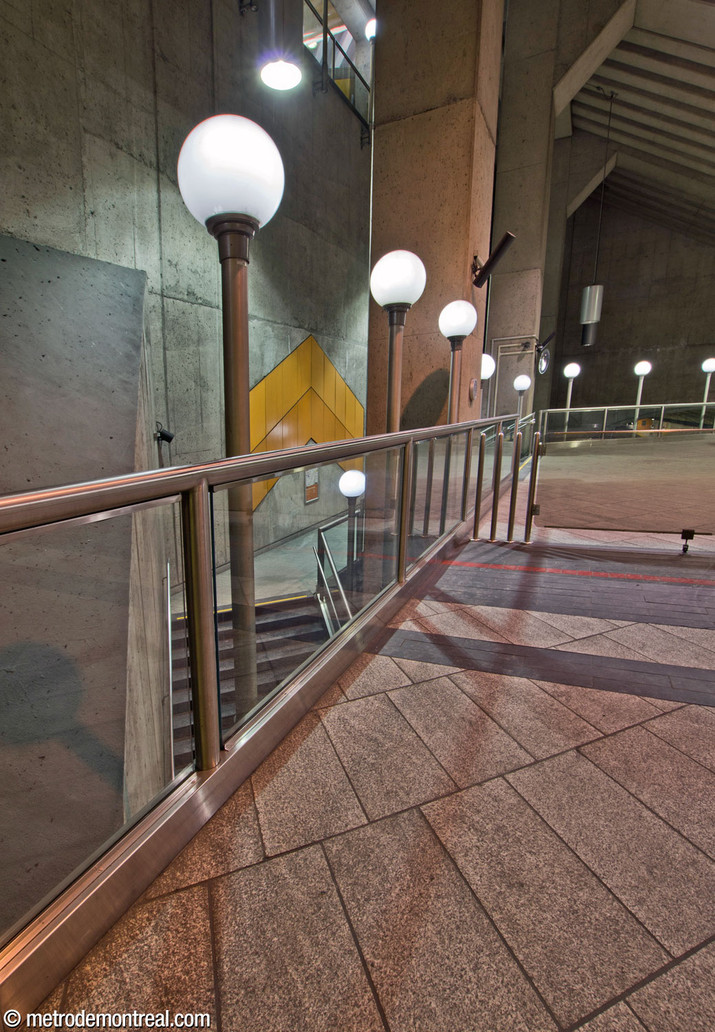 Another row of lamps lines the parapet along the eastern edge of the mezzanine, above the staircases to the Côte-Vertu platforms, which curl around the columns supporting the mezzanine and roof. A chevron by Gilbert Sauvé stands over the landing.