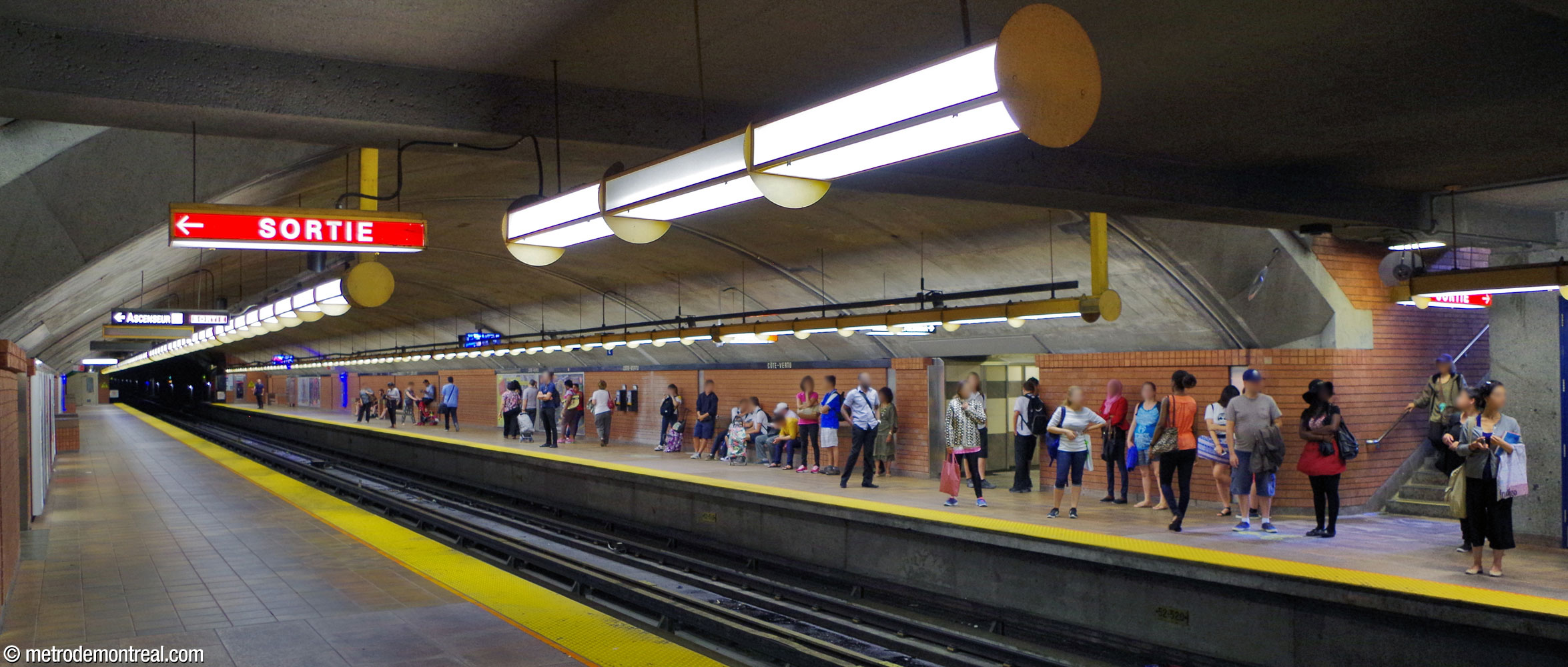 View of the busy departure platform. The niche for the elevator up to crossover level is located just left of the stairs. Note the distinctive yellow housings, end hangers, and circular plates that render the standard platform light fixtures unique.