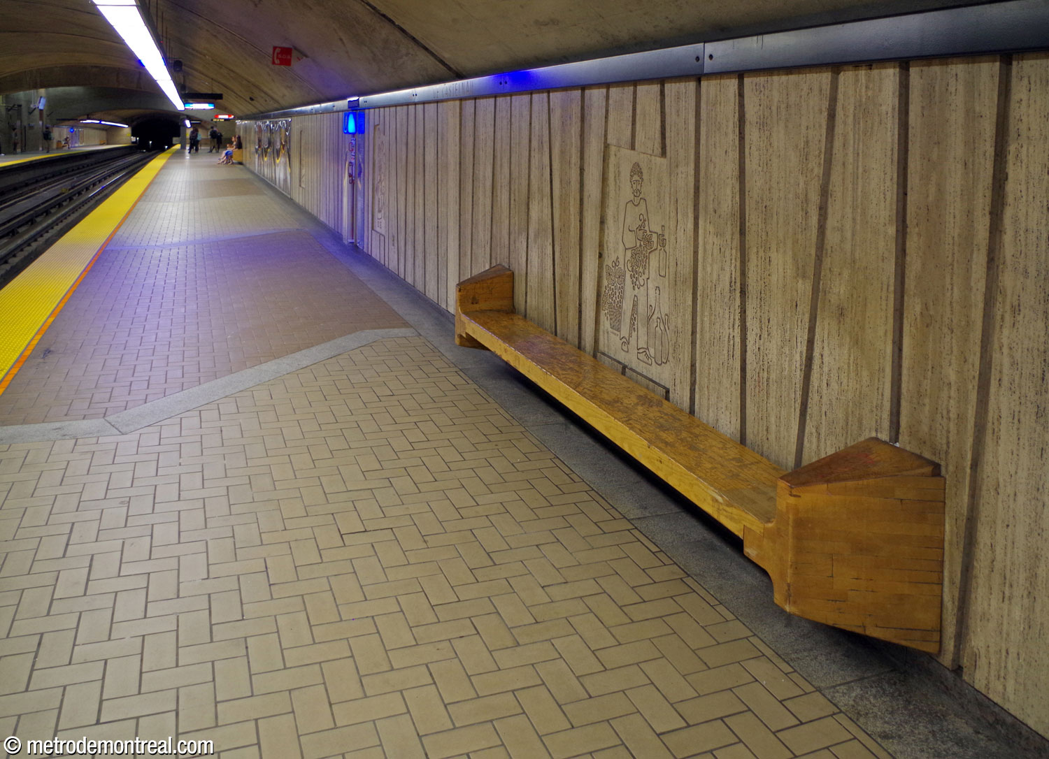 One of the wooden benches in the mined portion of the trainroom. Above the bench, the travertine wall panels frame a bas-relief by Jean-Charles Charuest. On the floor, the pattern of tan and brown floor tiles demarcated by grey stone is visible.