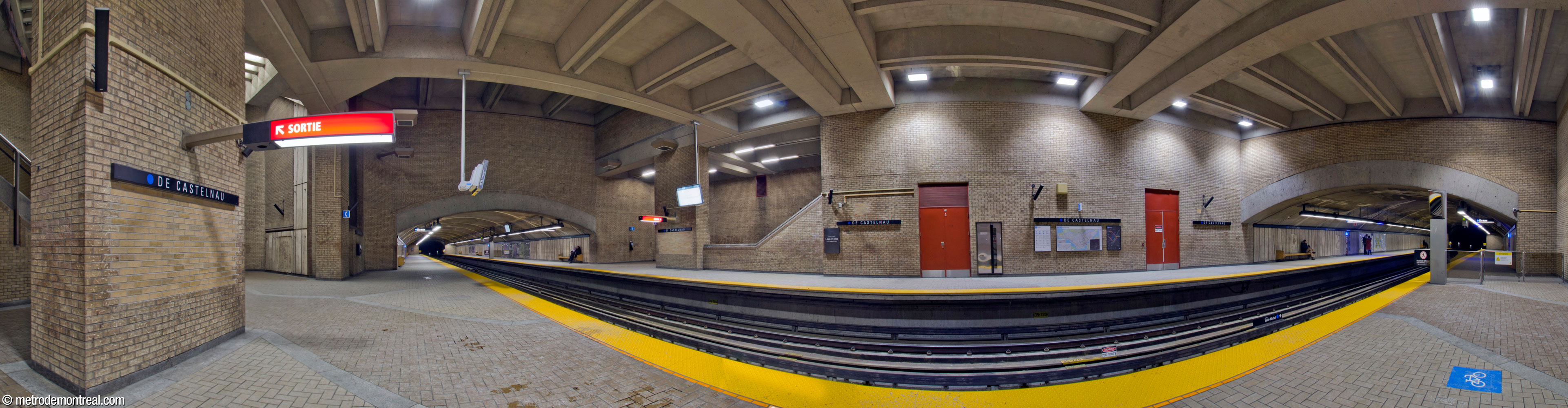 Panorama of the main volume of De Castelnau metro station, seen from the Saint-Michel platform. Large piers frame the staircases to the Saint-Michel (left) and Snowdon (centre) platforms. The mined trainroom sections extend to the east and west. The brickwork, floor tiling, and travertine panels form a consistent colour scheme of pale earth tones.