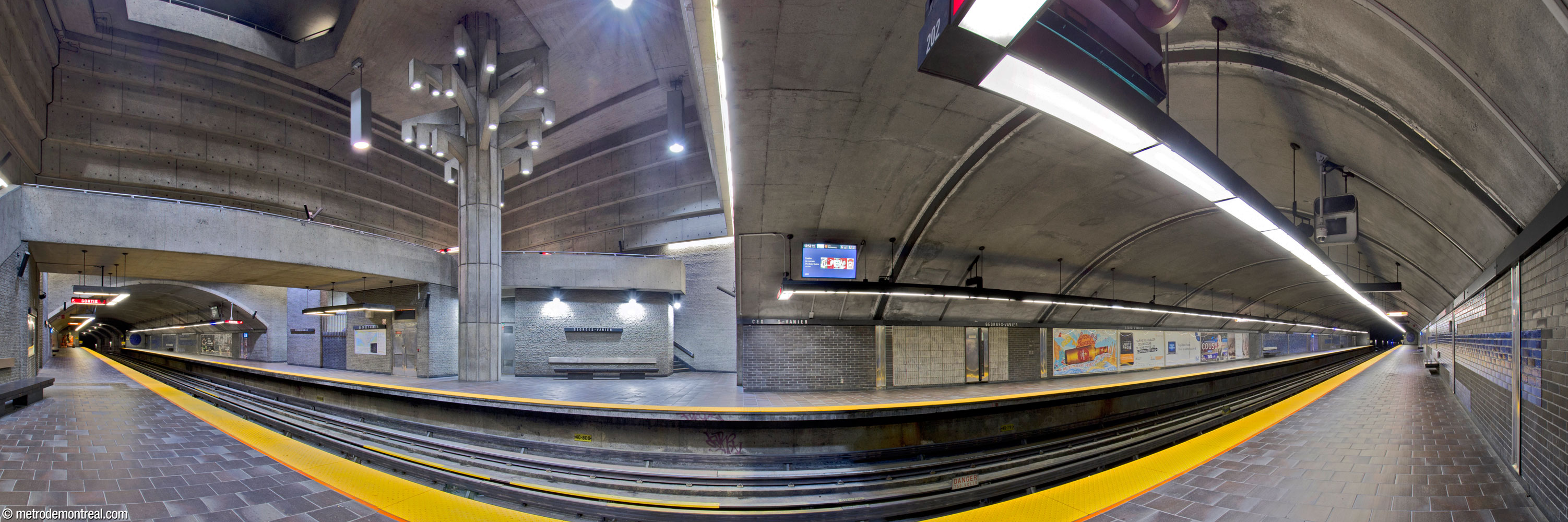 The trainroom of Georges-Vanier station. The tunnelled trainroom projects out on either side of the central volume, which contains a widened portion of each platform. The sculpture Un Arbre dans le parc rises up through the volume above the widened portion of the Côte-Vertu platform, in front of the crossover (left). The volume is clad in grey concrete bricks below and exposed concrete above; the portions of the platforms built in tunnel are clad in black and sapphire blue bricks.