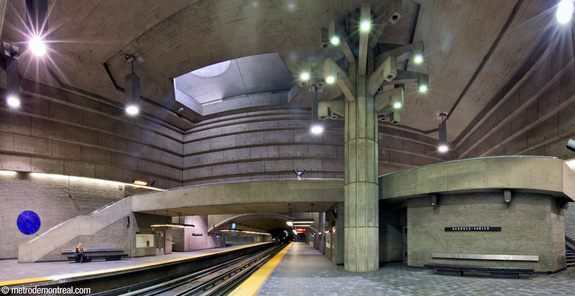 Panorama of the central volume and crossover, seen from the Côte-Vertu platform. Above the concrete brick lower walls, the upper part of the irregularly shaped volume features stepped concrete walls. The sculpture Un Arbre dans le parc rises up through the volume, towering over a wide seating area on the platform and another on the crossover above.