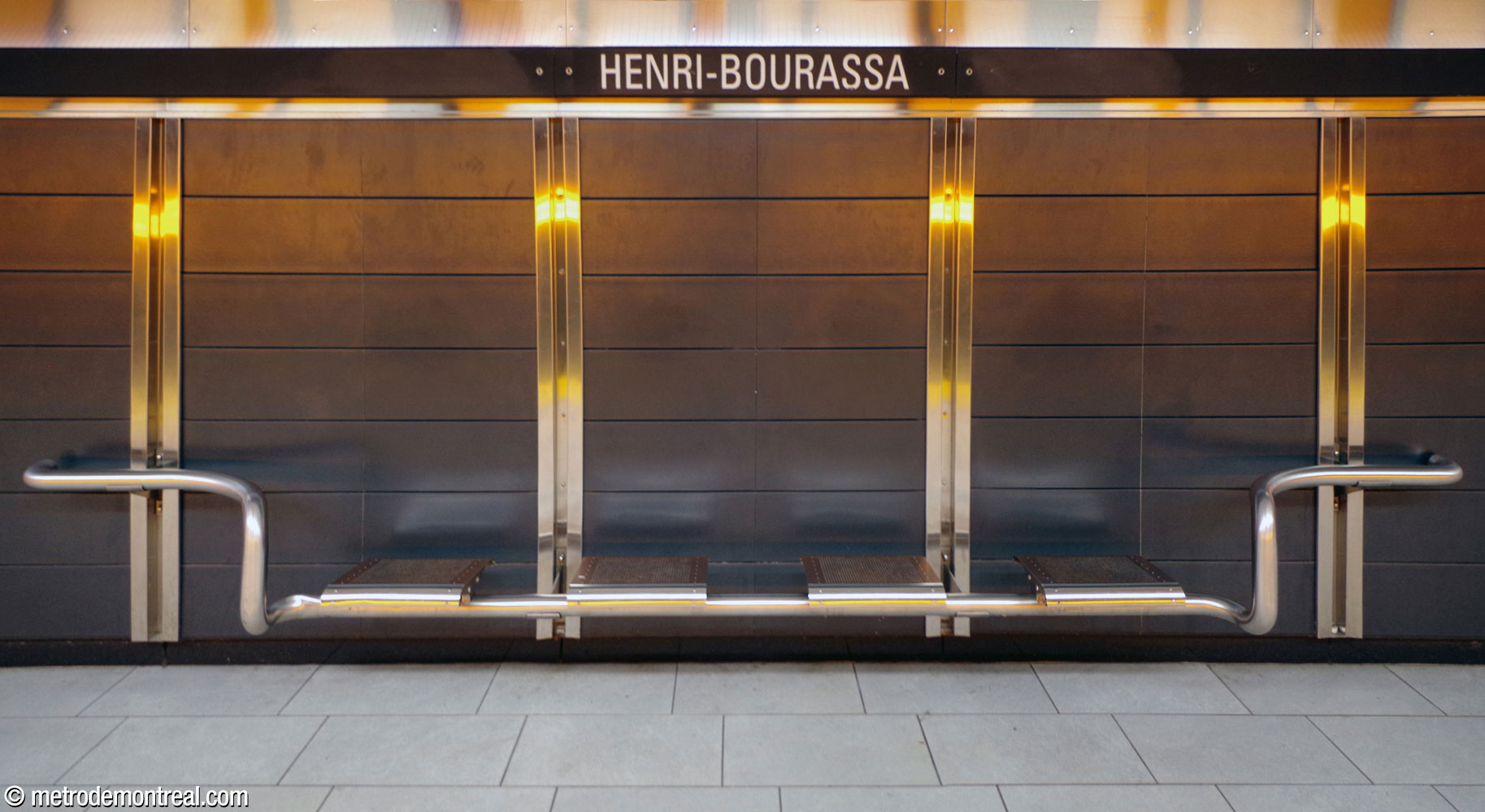 One of the stainless steel benches along the Montmorency platform, against the grey panels and stainless steel seams of the wall of the platform wall. The reflections from the orange lights above the tracks are also seen.