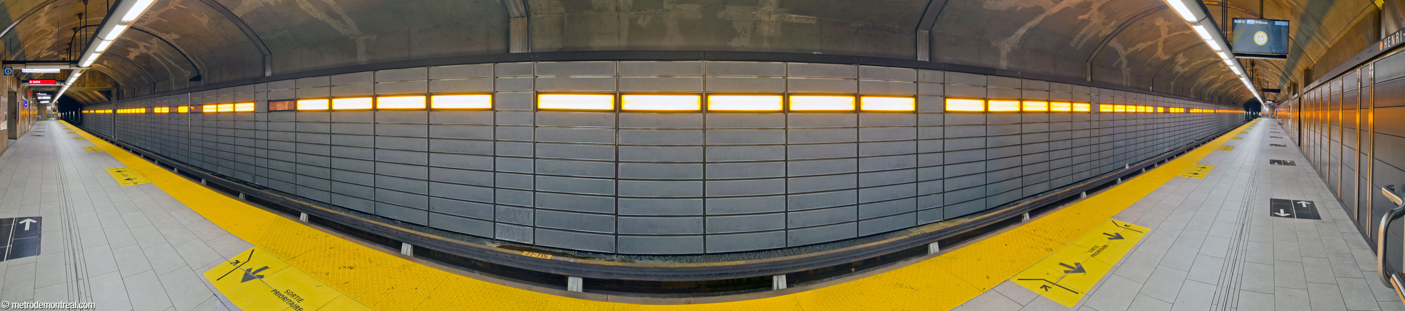 Panorama of the new trainroom, hosting the Montmorency platform. The walls are clad in slate-grey panels; the wall above the tracks hosts a strip of bright orange lights.