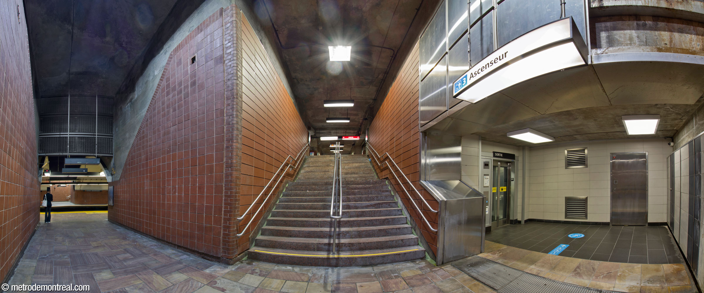 The north stairwell to the Côte-Vertu platform. Each of the four stairwells to the original trainroom turns a 90-degree corner as it leads to the platforms. The two northern stairwells descend all the way to platform level before turning the corner. The hallway from the foot of the stairs to the platform faces its counterpart on the other side, forming a box intersecting the tunnel vault. The upper part of the opening is now screened with a metal grille, presumably for fire safety. The elevator to the crossover level is at right.