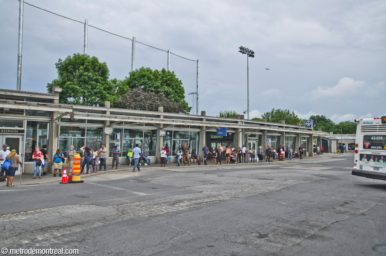 Crowds await buses outside the south building of the Henri-Bourassa Sud bus terminal, seen from outside the north building. The right-hand portion of the building contains the entrance to the station; it is separated from the enclosed jetty to the east by a short section that is roofed but open in front.