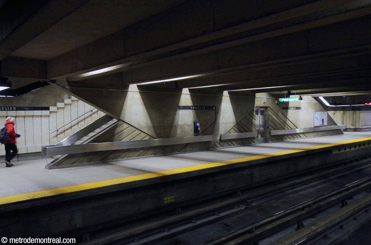 Vue de l'un des murs distinctifs des quais situés entre les escaliers vers la mezzanine, avec ses formes et reliefs anguleux et sa main courante en acier inoxydable en continuité avec celles des escaliers.