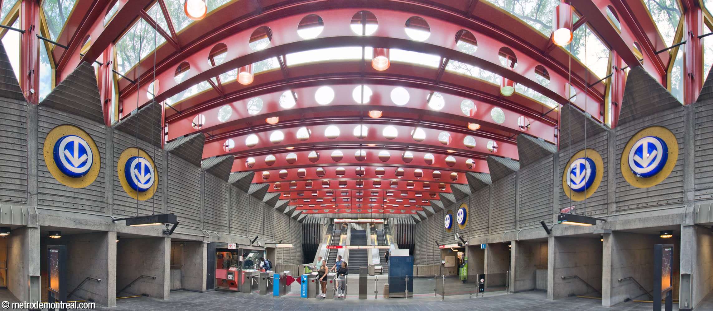 Vue d'ensemble de la mezzanine située dans le grand volume de la station, recouvert et illuminé par le plafond vitré de l'édicule sud. À l'extrémité, des escaliers se dirigent vers la sortie. Des logos du métro indiquent les escaliers vers les quais ainsi que le tunnel vers la sortie nord; chacun de ces éléments est articulé par une division en travées de largeur égale.