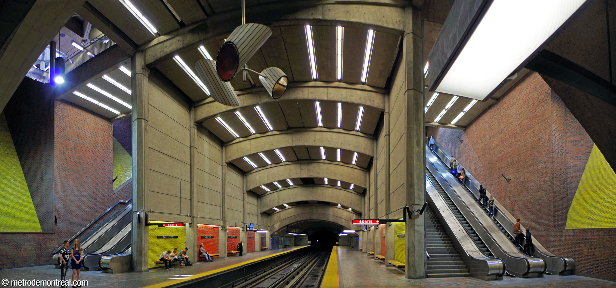 The station's main volume seen from the Côte-Vertu platform. The towering volume, receding into stepped vaults as it approaches the western end of the station, is separated by tall curtain walls from the deep escalator shafts on either side. Jacques de Tonnancour's sculpture is suspended from the ceiling.