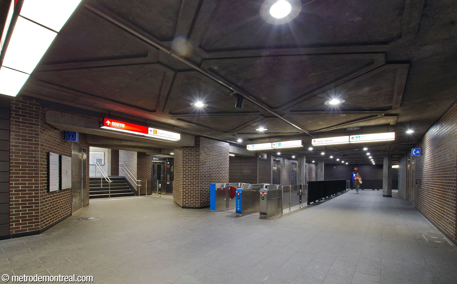 The hallway toward the two eastern headhouses. A stairwell, left, heads up to the Saint-Jacques exit; the hallway continues at right toward the Place Saint-Henri exit. At centre, a gateline leads into the fare-paid zone that connects the station’s two elevators. The elevator to the Côte-Vertu platform is at centre-left; the fare-paid zone runs along the hallway to reach the second one.