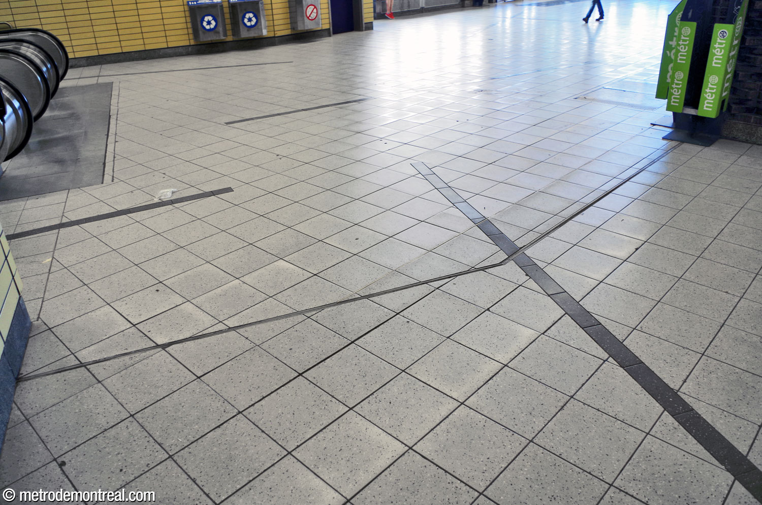 Le plancher de l'accès Berri Ouest. Des lignes obliques de carreaux gris foncé interrompent la régularité du carrelage pâle; ce motif relie l'ensemble des niveaux supérieurs de la station.