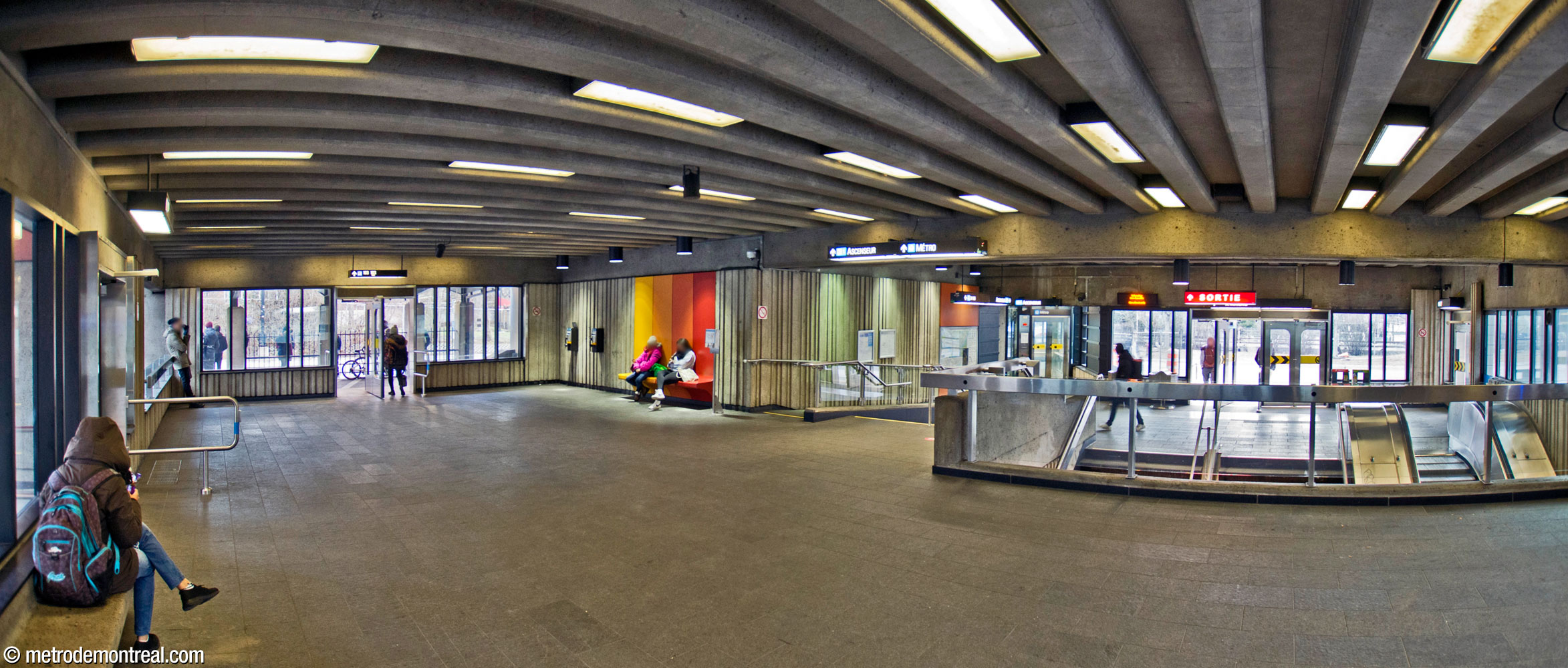 The interior of the headhouse seen from the raised rear section. The elevator is visible in the background at centre-right, next to the main exit. The row of seats at centre-left shares the same colour scheme as those on the platform.