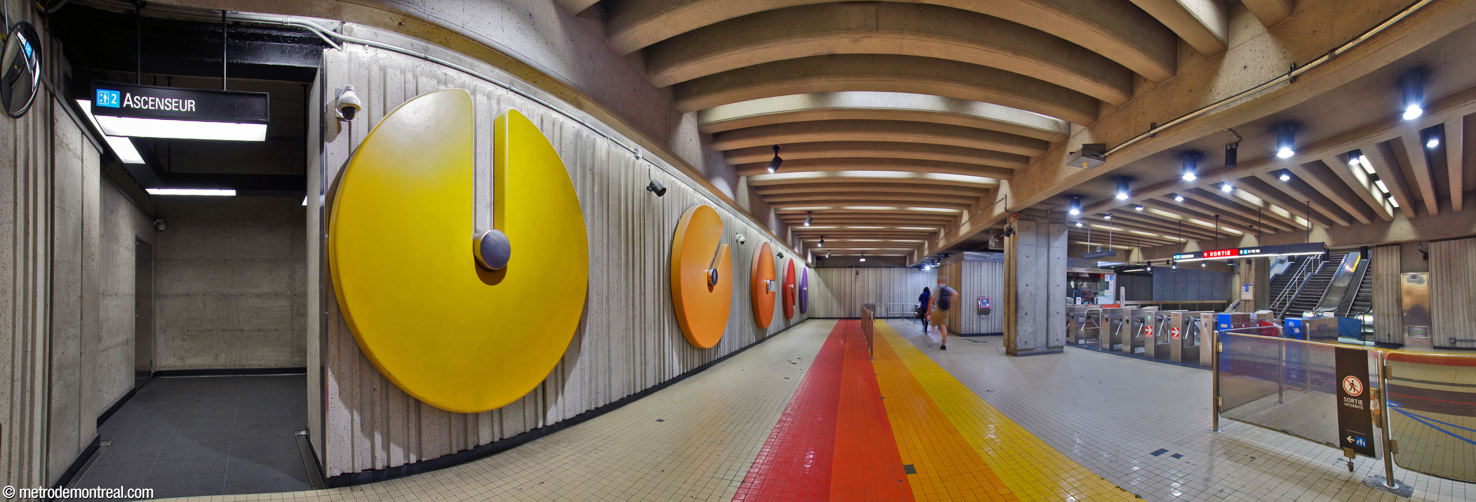 Panorama of the ticket hall seen from the northeast corner. The head of the escalators to transept level is at centre-right; at far left is the entrance to the elevator down to the transept and the Côte-Vertu platform. Unusually, a line of barriers separates entering from exiting passengers, with exiting passengers passing along the back wall. At left, five mural sculptures by André Léonard ornament the back wall. The pale tiling of the floor is interrupted by bright stripes of red, red-orange, orange, and yellow, repeating the colours of the sculptures and the designs at platform level.