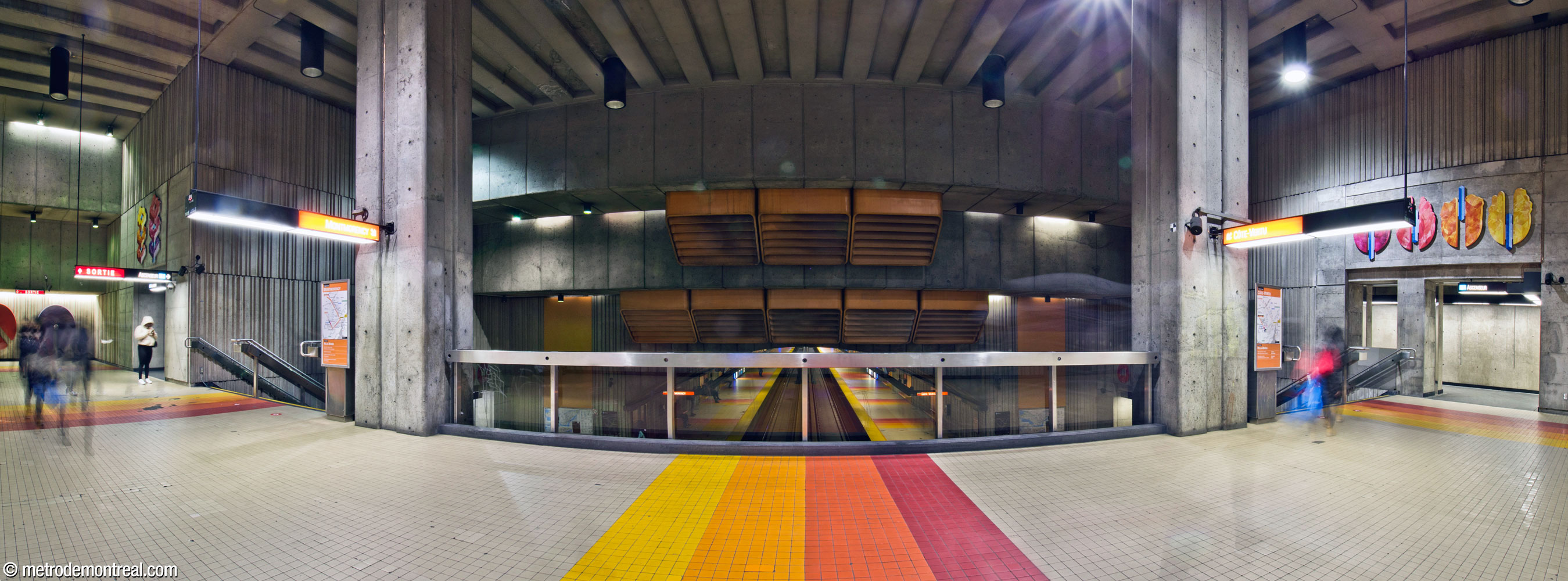 The transept level, crossing over the tunnel mouth at the south end of the platforms. The high ceiling descends toward the platforms in a series of steps over the north wall, hosting an array of large orange ventilation outlets framed by the imposing pillars supporting the ceiling. Mural sculptures by Marianne Chevalier are seen over the elevator entrances at the right and left.