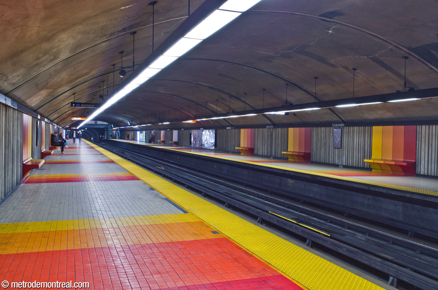 View of the station's trainroom, dug in tunnel, looking south from the Côte-Vertu platform. Each bench module features a gradient of yellow, orange, red-orange, and red, accompanied by matching stripes on the floor; the yellow colour marks the side closest to daylight, that is, to the exit.