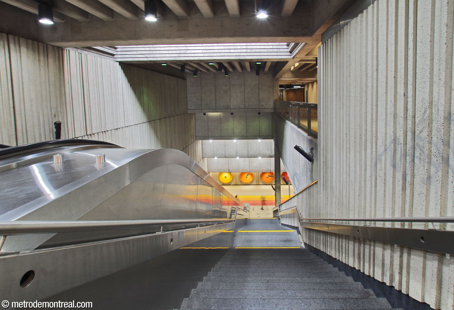 The volume of the main stairwell between the ticket hall and the transept level. A light shaft above provides natural light. At top right, the walkway from the upper elevator to the ticket hall overlooks the volume. The second suite of mural sculptures by André Léonard decorates the wall facing the foot of the escalators.